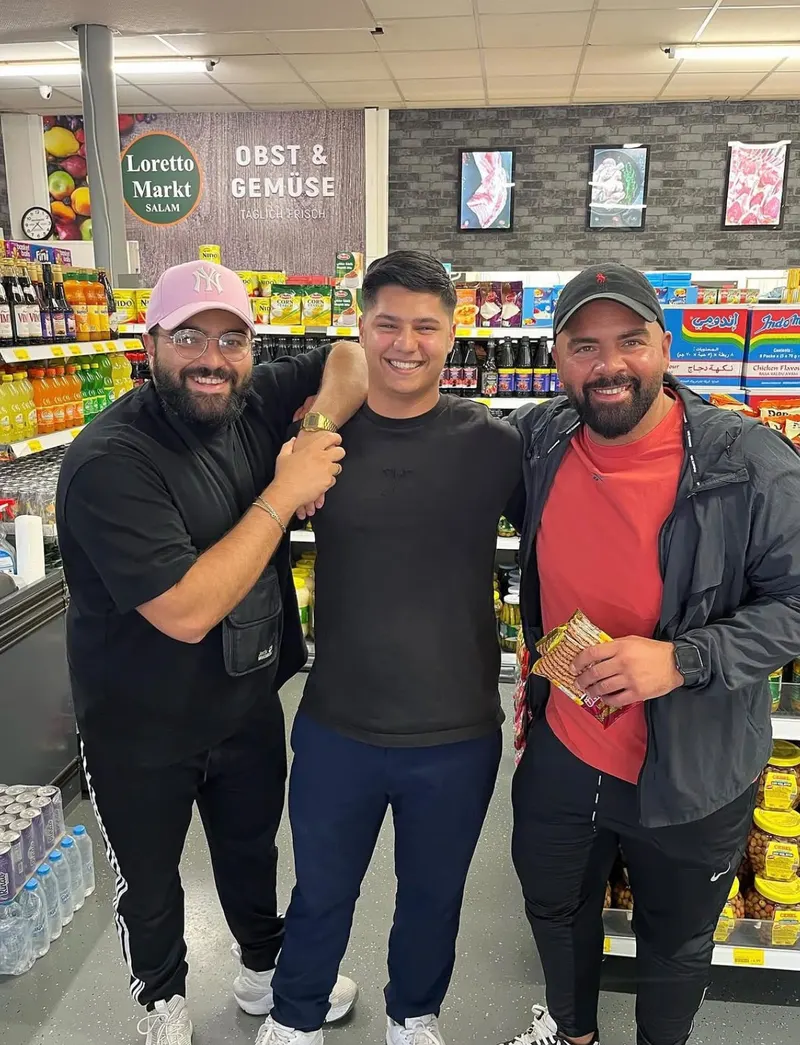 Serdar Karibik und Abdel Boudi Bassam „The Butcher“ lachend mit einem jungen Mann im Loretto Markt Salam, im Hintergrund das Schild Obst & Gemüse